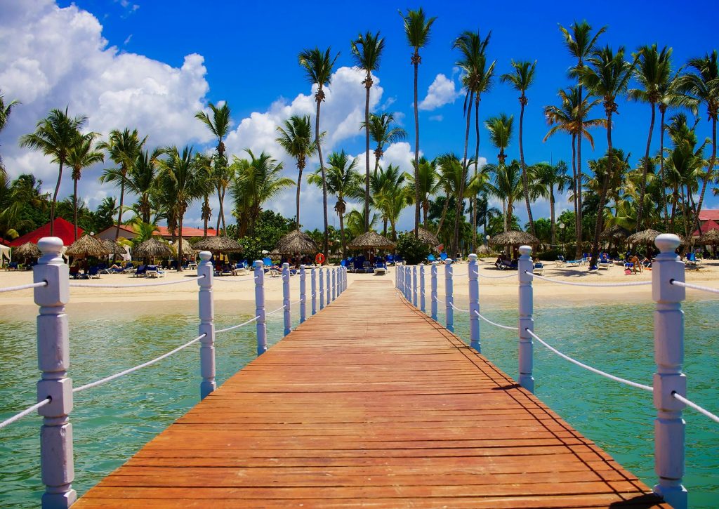 Wooden dock leading to a tropical beach resort with palm trees and clear blue waters.