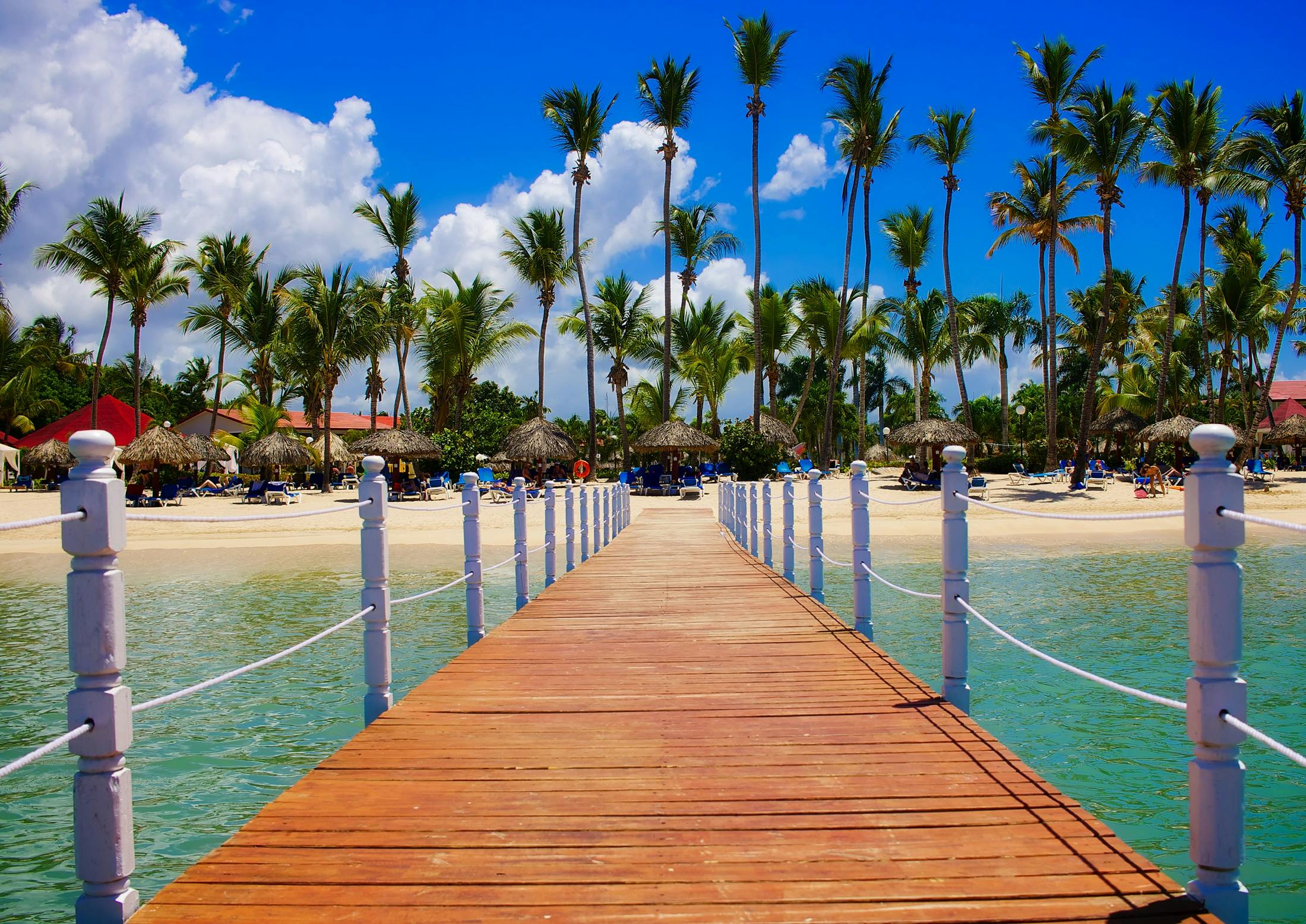 why-choose-us Wooden dock leading to a tropical beach resort with palm trees and clear blue waters.